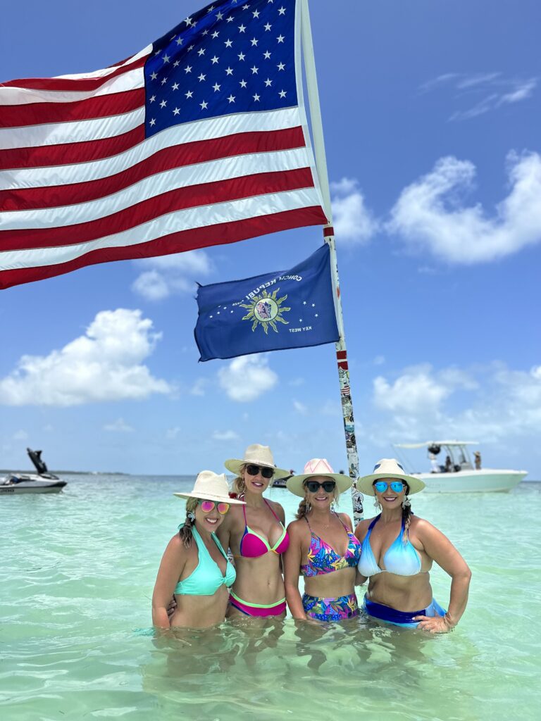 Four women standing in shallow waters at Islamorada Sandbar, Florida, with an American flag waving in the background